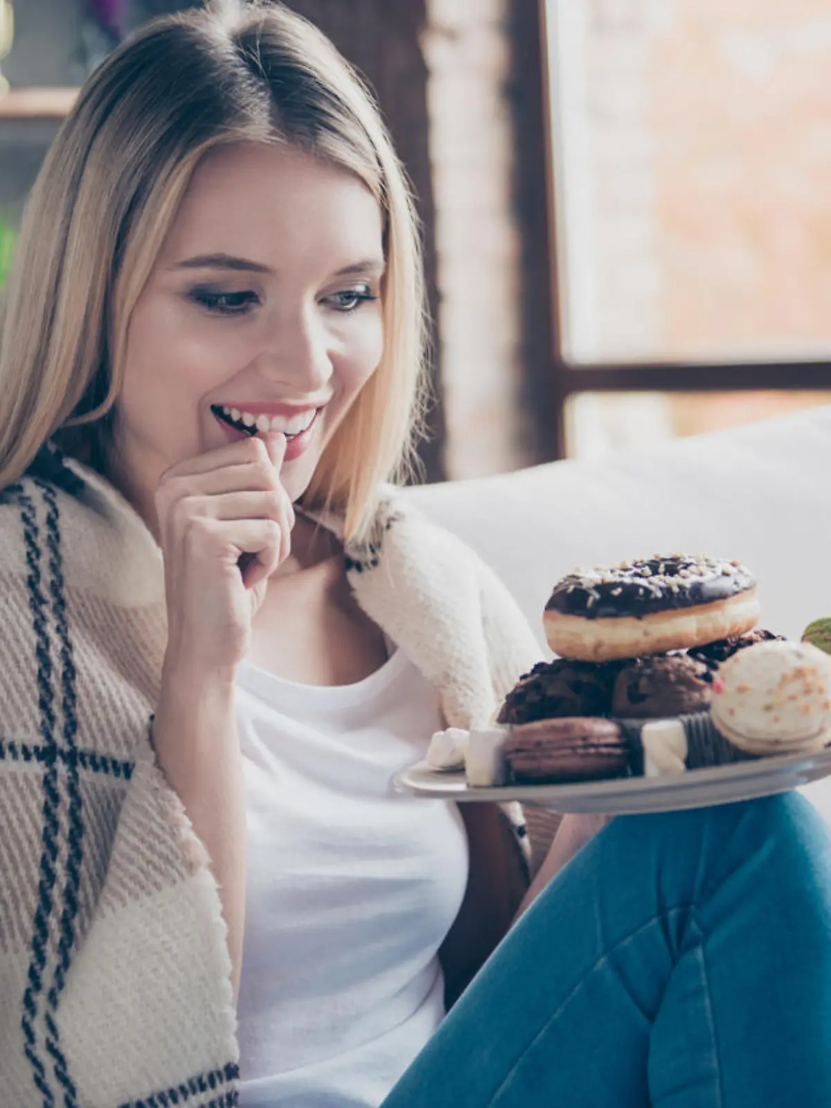 Portrait of beautiful emotional charming attractive sweet toothy woman sitting on sofa in living room, holding plate of donuts and macaroons, looking exciting satisfied