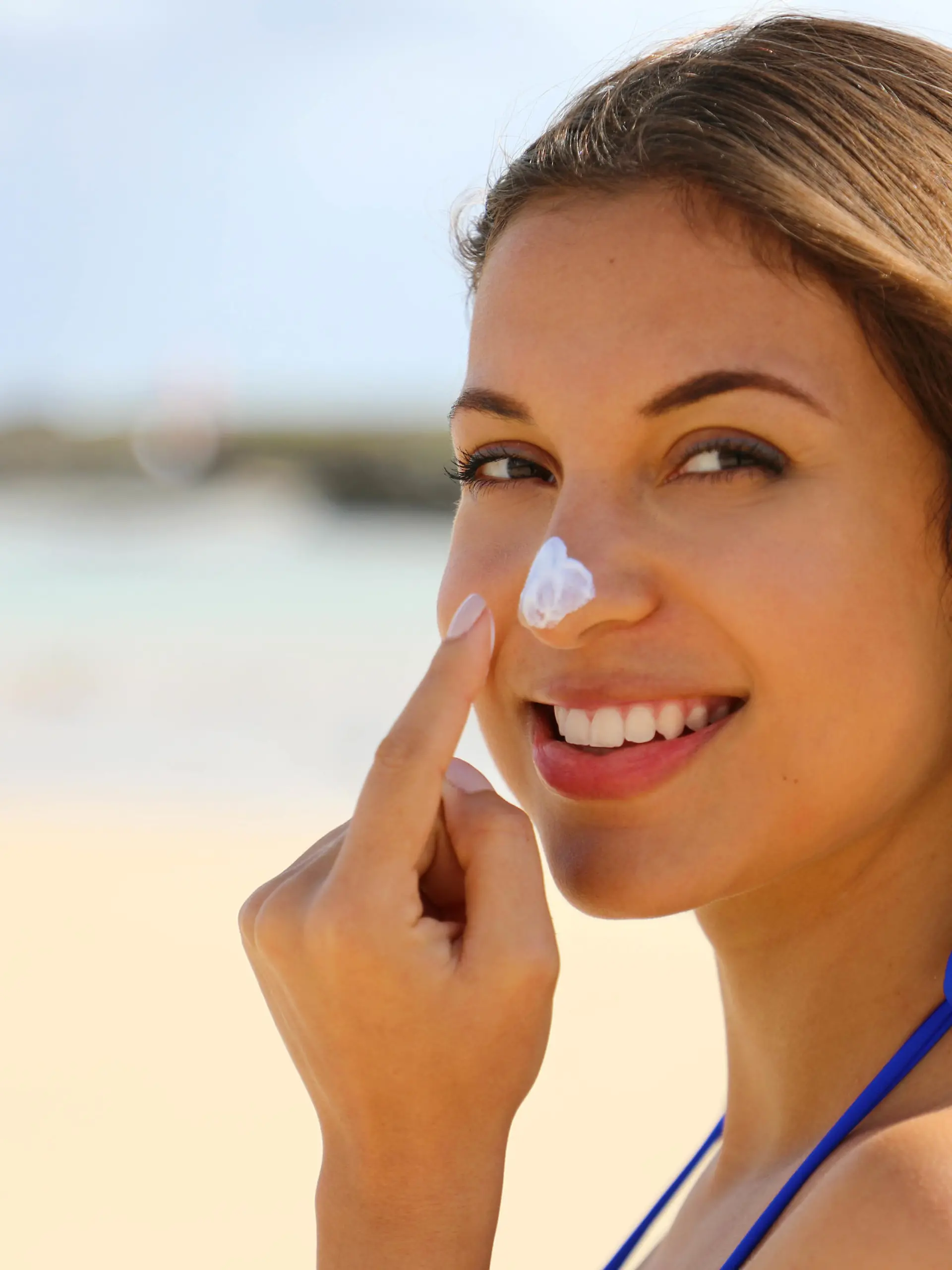 Close up young beautiful woman applying sun cream protection on her nose on the beach. Suntan lotion woman applying sunscreen solar cream.