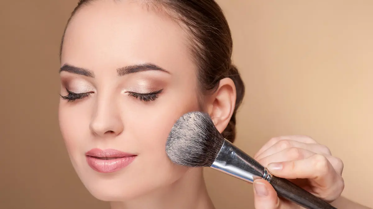 Close up of hand of professional make-up artist applying powder on female check. She is holding a brush. The woman closed eyes with pretty smile