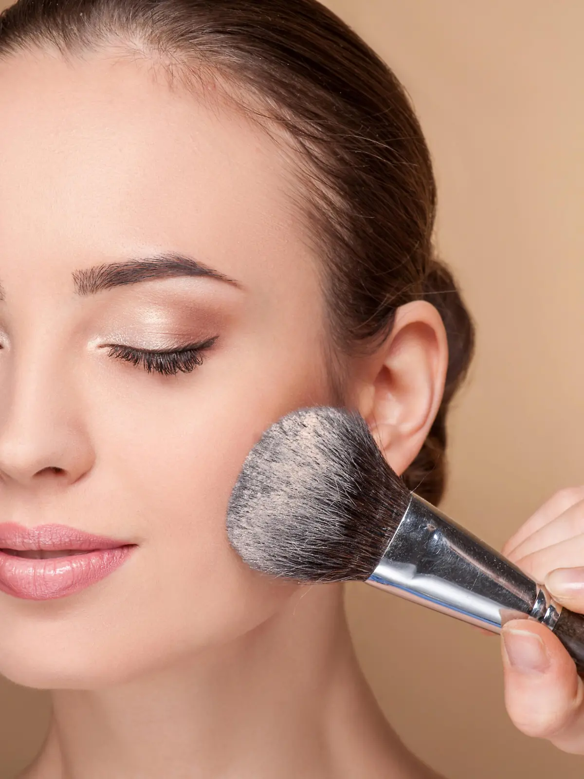 Close up of hand of professional make-up artist applying powder on female check. She is holding a brush. The woman closed eyes with pretty smile