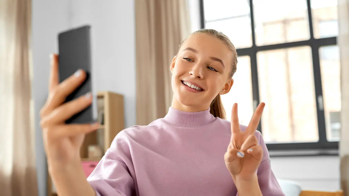 people, children and technology concept - happy smiling teenage girl with smartphone taking selfie and showing peace gesture at home || Modellfreigabe vorhanden