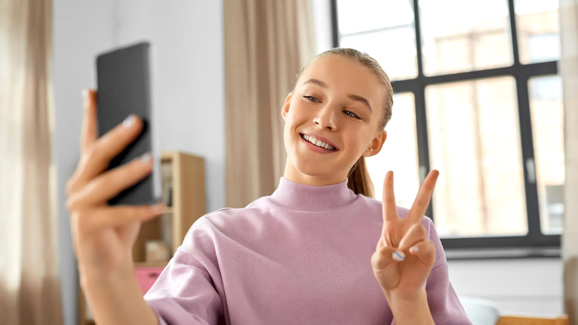 people, children and technology concept - happy smiling teenage girl with smartphone taking selfie and showing peace gesture at home || Modellfreigabe vorhanden