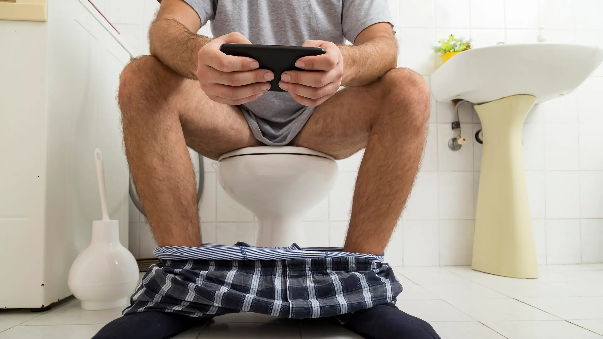 Detail of a man sitting on the toilet, reading the newspaper on a smart phone