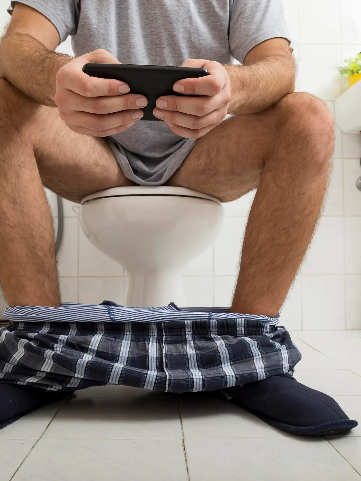 Detail of a man sitting on the toilet, reading the newspaper on a smart phone