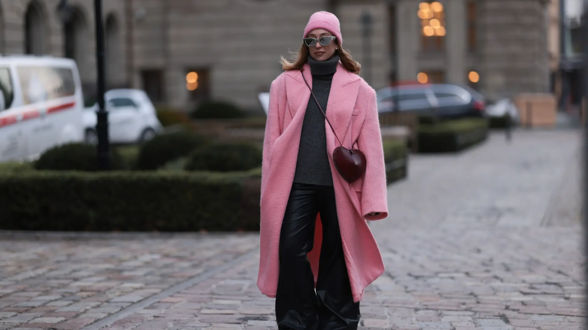 Influencerin Sonia Lyson trägt zu ihrer weiten schwarzen Hose einen dunkelgrauen Rollkragenpullover und kombiniert eine pinke Mütze und einen pinken Mantel