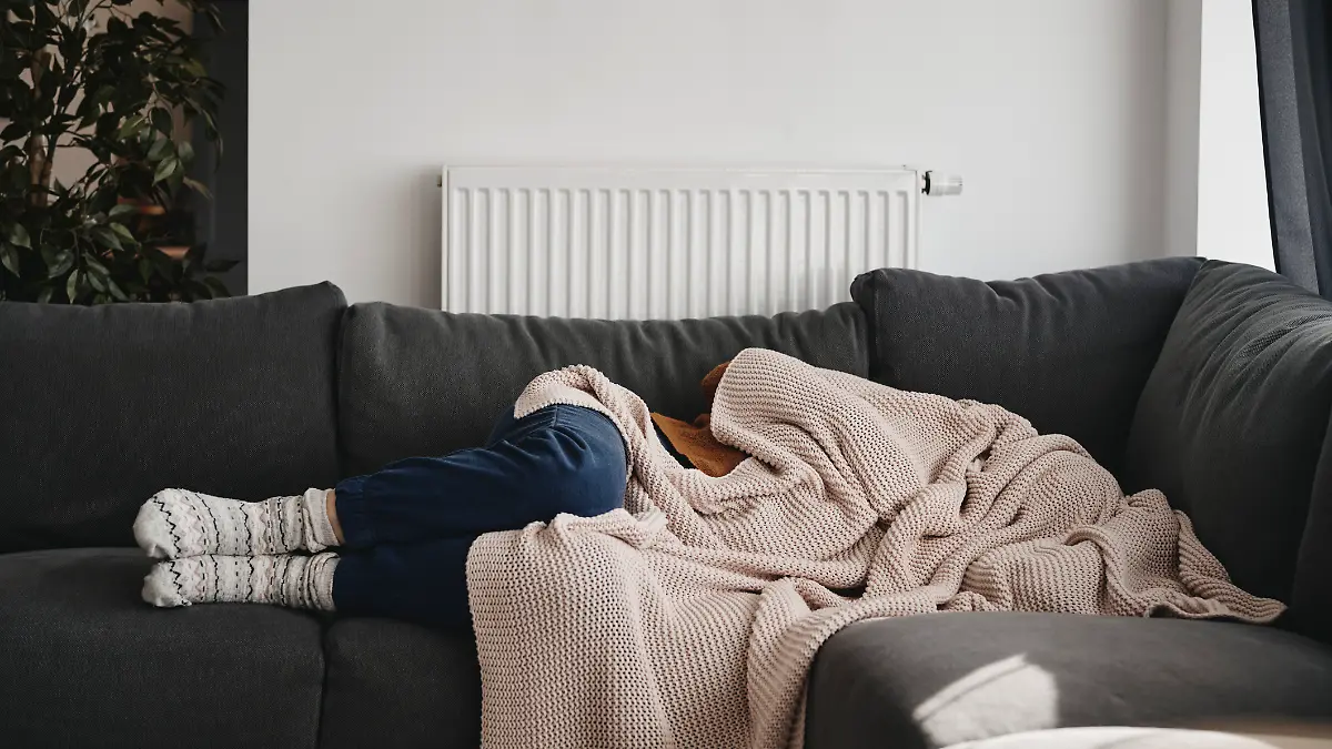 Eine Frau liegt krank oder traurig auf der Couch und hat sich die Decke über den Kopf gezogen.