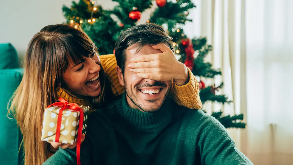 Eine Frau hält einem Mann die Augen zu und überrascht ihn mit einem Geschenk an Weihnachten.