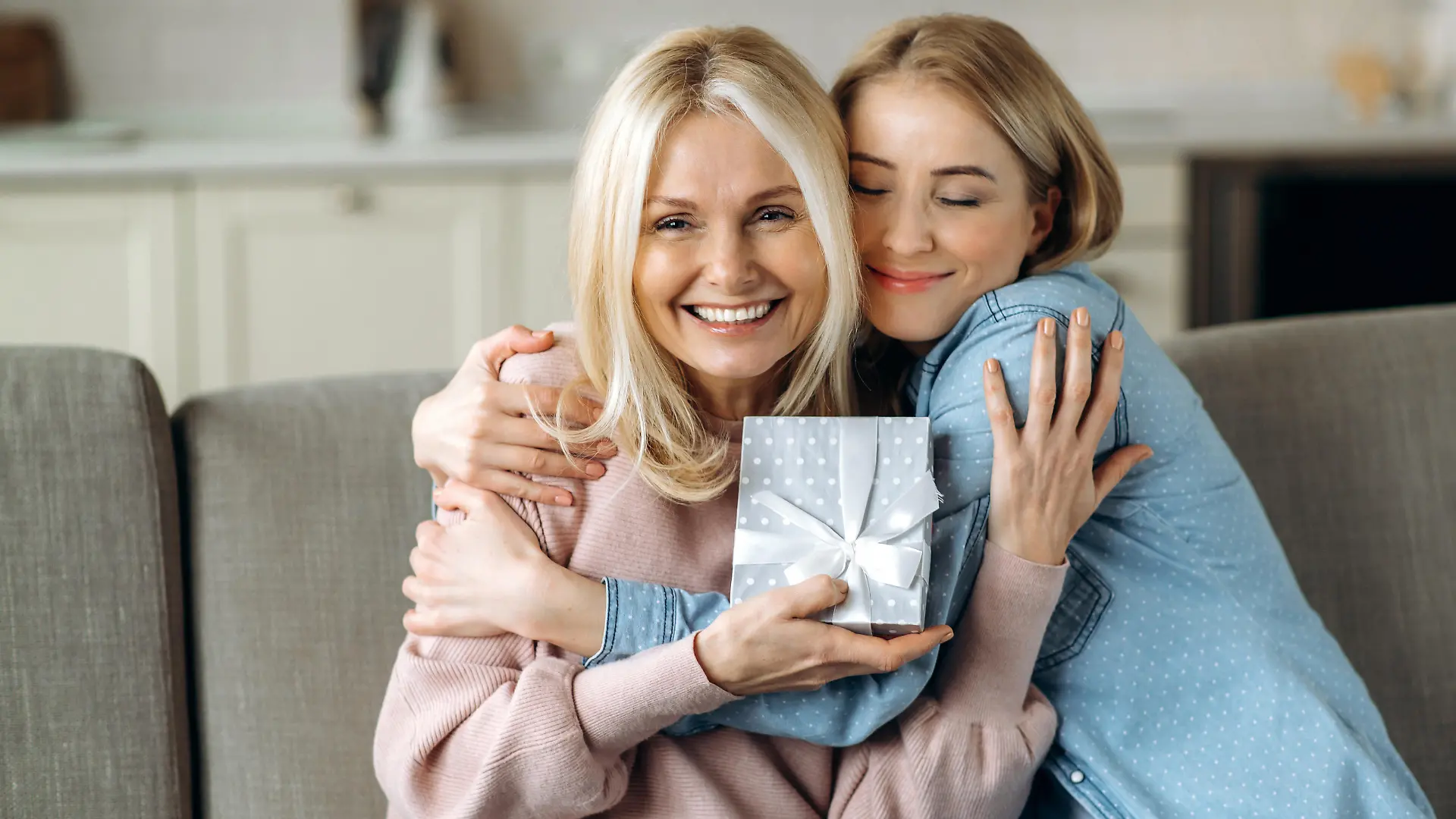 Surprise for mother's day or birthday. Loving young adult daughter giving a present to her beloved middle-aged caucasian mom sitting on the sofa in the living room