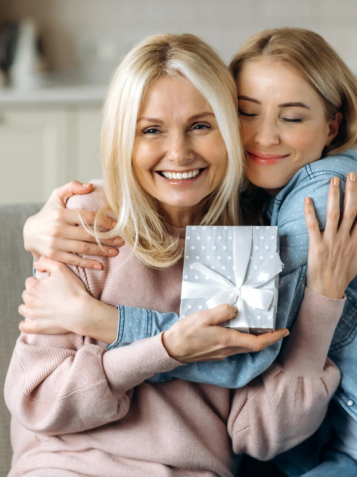 Surprise for mother's day or birthday. Loving young adult daughter giving a present to her beloved middle-aged caucasian mom sitting on the sofa in the living room