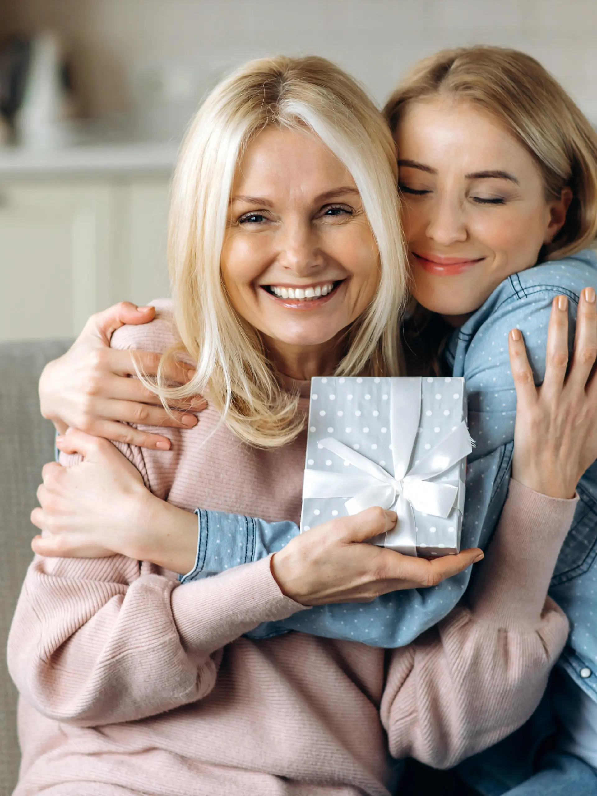Surprise for mother's day or birthday. Loving young adult daughter giving a present to her beloved middle-aged caucasian mom sitting on the sofa in the living room