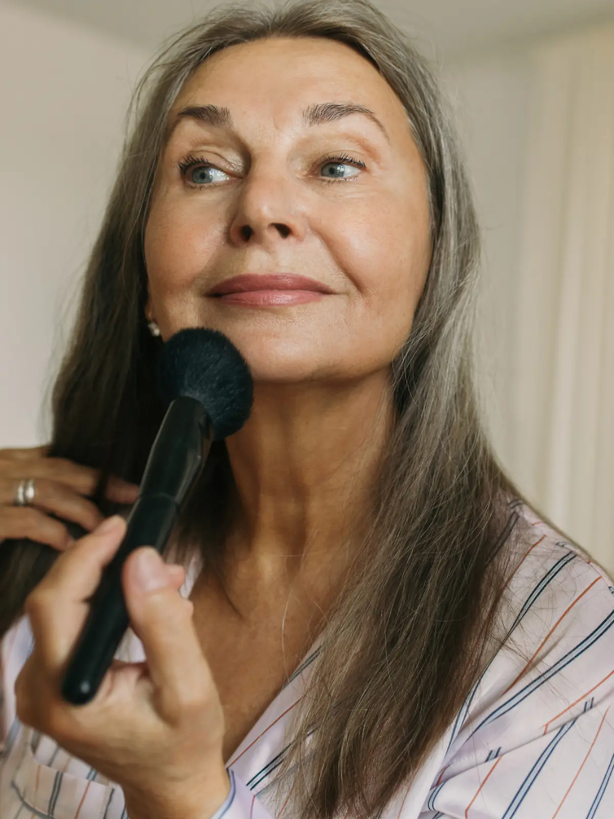 Eine ältere Frau steht vor dem Spiegel und trägt mit einem Pinsel Make-up auf.