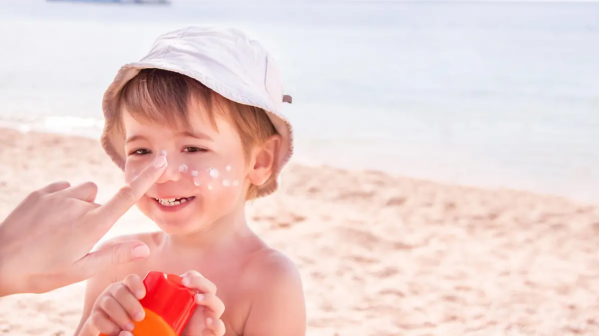 Sun protection. Hand of caucasian mother applying suncream  (suntan lotion) from a plastic container to her happy cute son before tanning during summer holiday on beach. Summer vacations concept. Copyspace, close up.