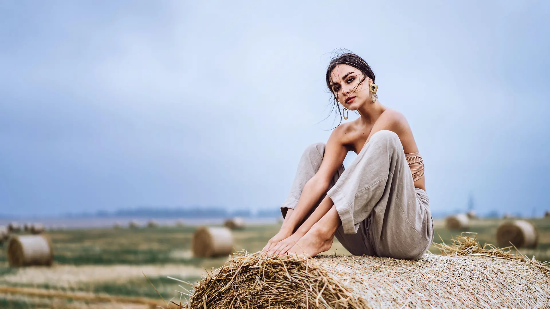 Brunette in linen pants and bare shoulders sitting on a hay bales in warm autumn day. Woman looking at camera. Behind her is a wheat field.