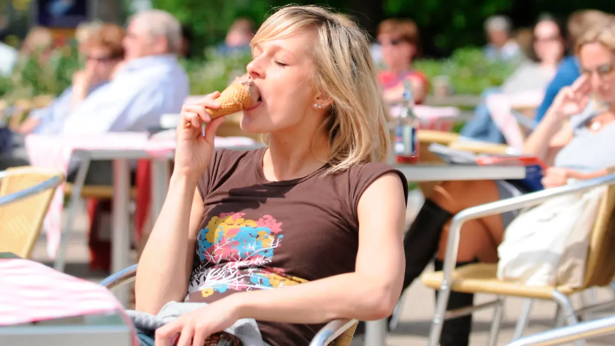 Junge blonde Frau sitzt in einem Café im Sonnenschein, isst ein Eis, sonnt sich, entspannt || Modellfreigabe vorhanden