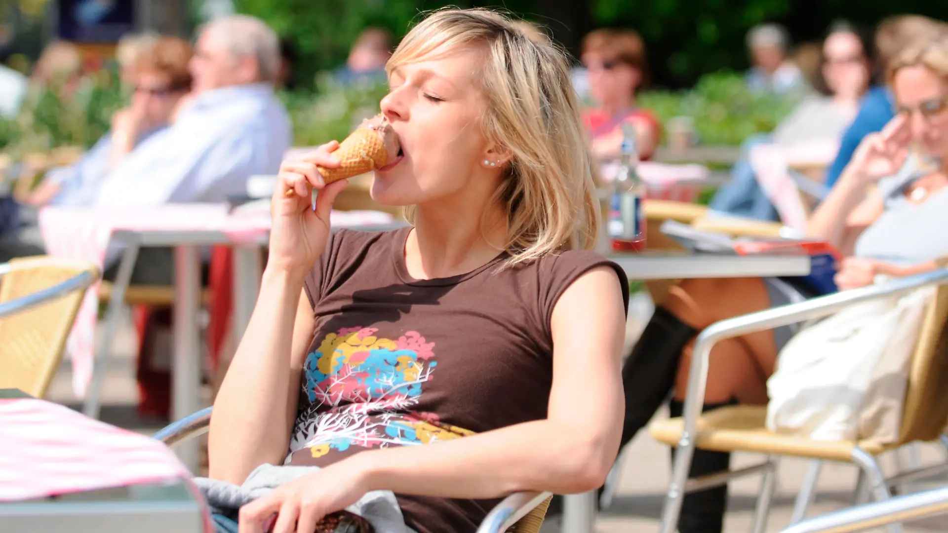 Junge blonde Frau sitzt in einem Café im Sonnenschein, isst ein Eis, sonnt sich, entspannt || Modellfreigabe vorhanden