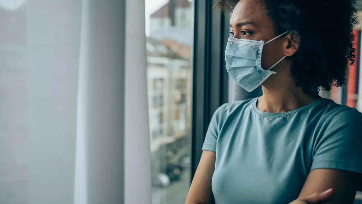 Beautiful african-american woman wearing protective face mask and looking through window at home during Coronavirus/COVID-19 pandemic.