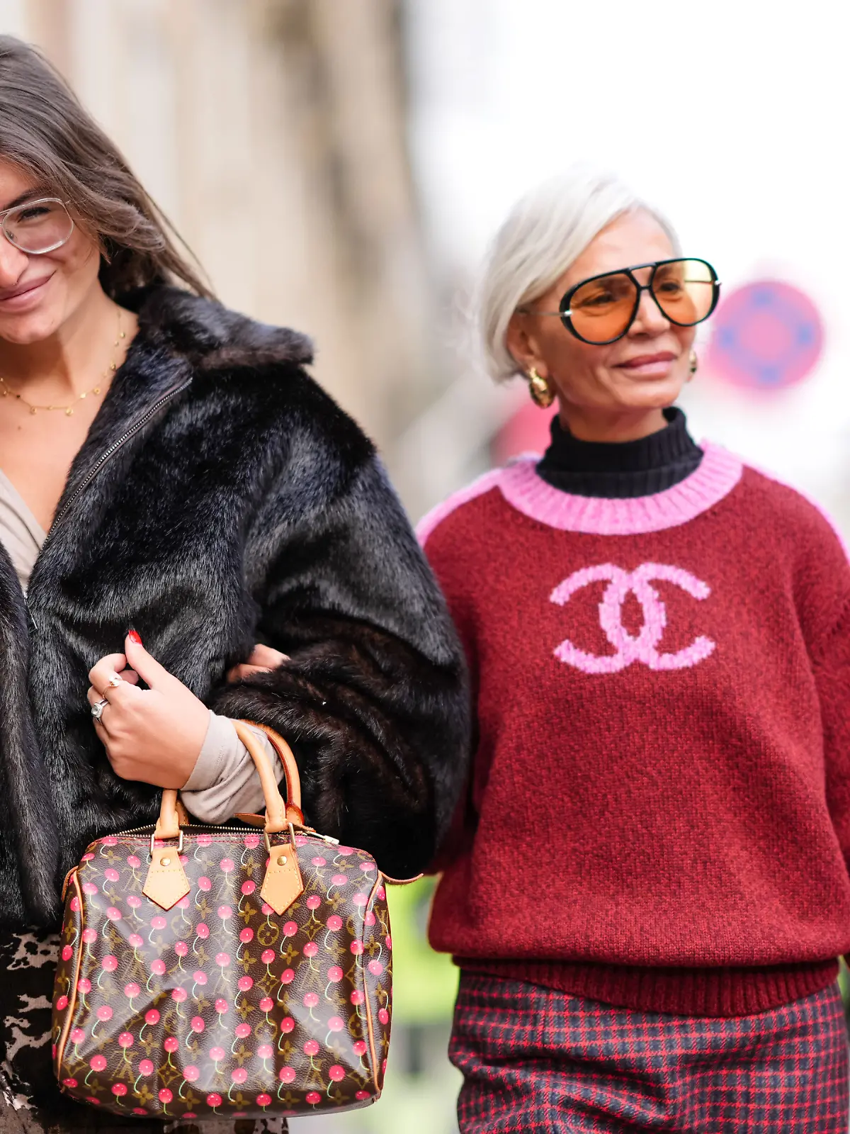 Eine braune Vuitton-Ledertasche mit Monogramm-Logo und aufgedruckten Kirschen, einen Minirock, schwarze Strumpfhosen und schwarze spitze Schuhe; Grece Ghanem (R) trägt eine Sonnenbrille, goldene Ohrringe, einen schwarzen Rollkragenpullover und ein rosa-roter Wollstrickpullover mit Chanel-Logo.