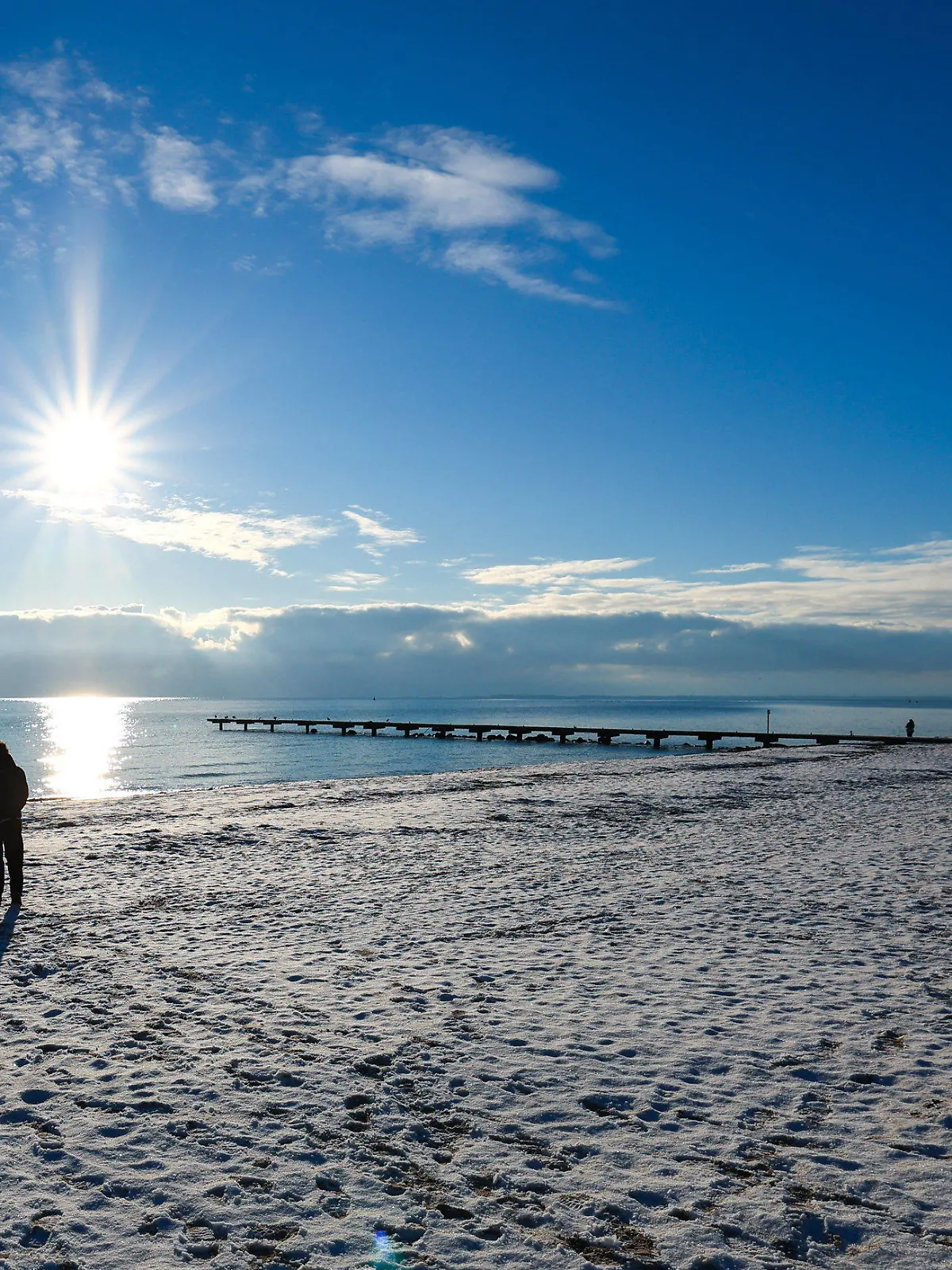ARCHIV - 16.12.2022, Schleswig-Holstein, Burg: Drei Menschen gehen am Südstrand bei Burg auf Fehmarn im strahlenden Sonnenschein spazieren. Der Sandstrand ist mit einer dünnen Schneedecke bedeckt. Die Ferienorte Schleswig-Holsteins sind über die Feiertage und den Jahreswechsel wieder gut gebucht. (zu dpa: «Schleswig-Holstein zu Weihnachten und Silvester sehr nachgefragt») Foto: Christian Charisius/dpa +++ dpa-Bildfunk +++