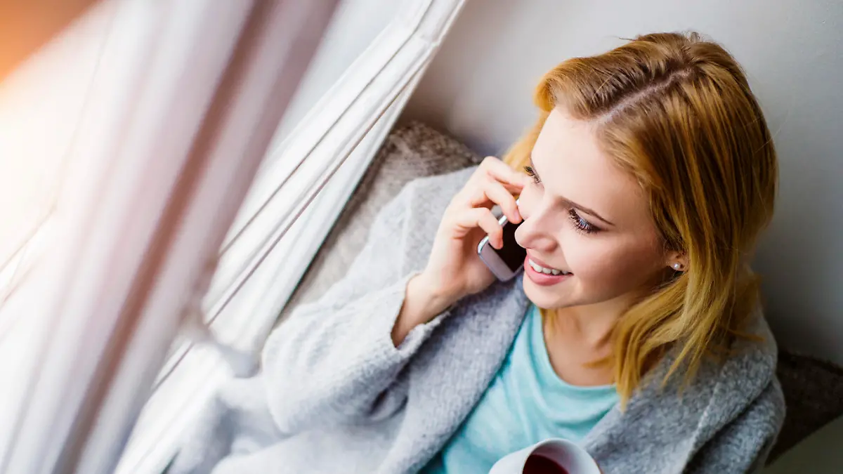 Beautiful blond woman sitting on window sill with smart phone and cup of tea making a phone call