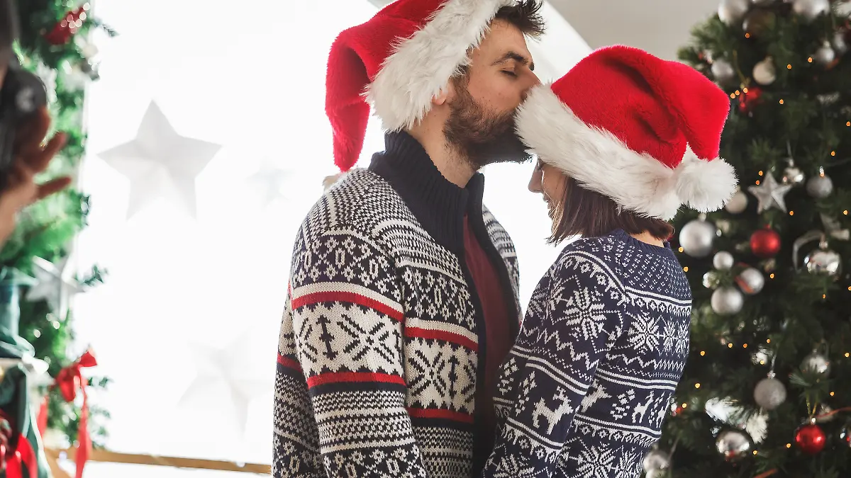 Ein Paar an Weihnachten: Der Mann küsst die Frau, die ebenfalls eine Weihnachtsmütze trägt, romantisch auf die Stirn.