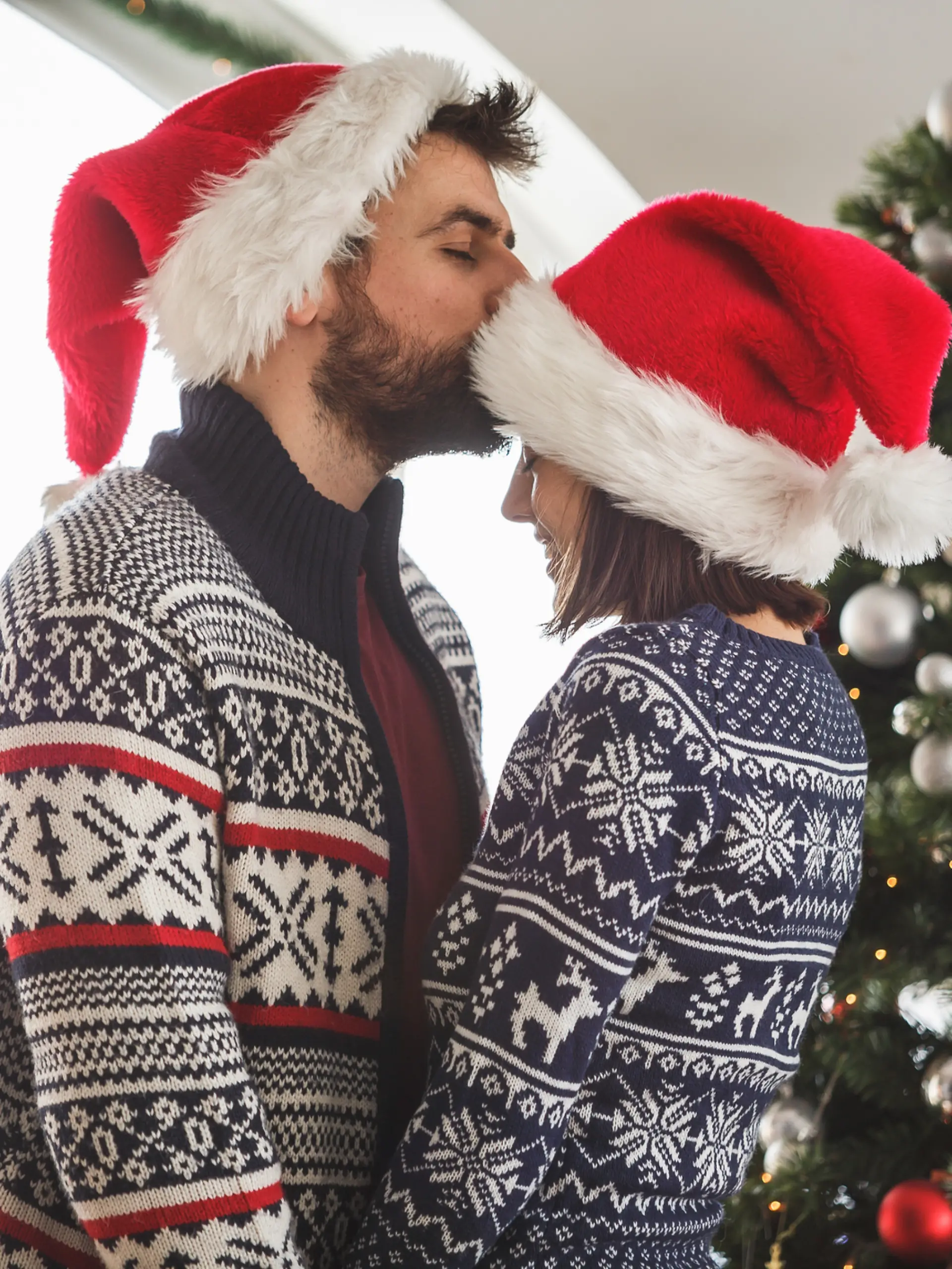 Ein Paar an Weihnachten: Der Mann küsst die Frau, die ebenfalls eine Weihnachtsmütze trägt, romantisch auf die Stirn.