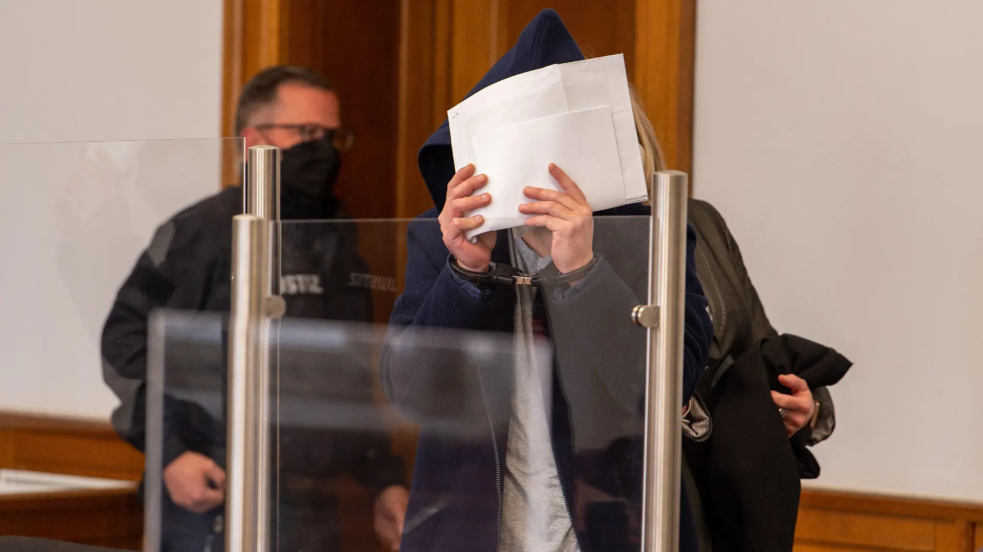 22.03.2022, Niedersachsen, Lüneburg: Der Angeklagte wird in den Saal vom Landgericht geführt und verdeckt sein Gesicht. Dem 38-Jährigen wird vorgeworfen, im Oktober 2021 in Lüneburg seine im 7. Monat schwangere Lebensgefährtin durch Messerstiche getötet zu haben. Foto: Philipp Schulze/dpa +++ dpa-Bildfunk +++