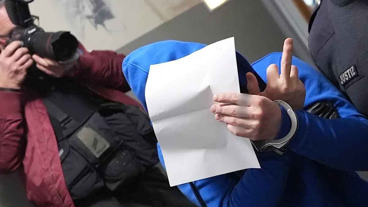 Schleswig-Holstein, Flensburg: Ein Angeklagter hält sich ein Blatt Papier vor sein Gesicht und zeigt dabei den Mittelfinger seiner rechten Hand auf dem Flur des Landgerichts, nach dem ersten Sitzungstag im Mordprozess. Vier Angeklagte sollen gemeinsam am 11. Januar 2024 ein Seniorenpaar in St. Peter-Ording brutal in dessen Haus überfallen haben.