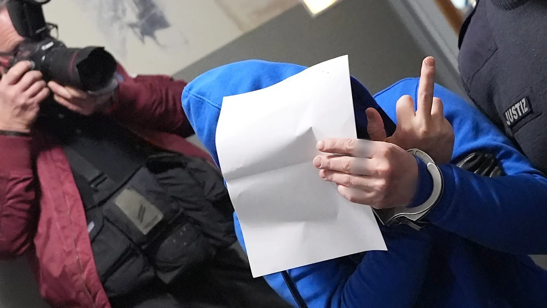 Schleswig-Holstein, Flensburg: Ein Angeklagter hält sich ein Blatt Papier vor sein Gesicht und zeigt dabei den Mittelfinger seiner rechten Hand auf dem Flur des Landgerichts, nach dem ersten Sitzungstag im Mordprozess. Vier Angeklagte sollen gemeinsam am 11. Januar 2024 ein Seniorenpaar in St. Peter-Ording brutal in dessen Haus überfallen haben.