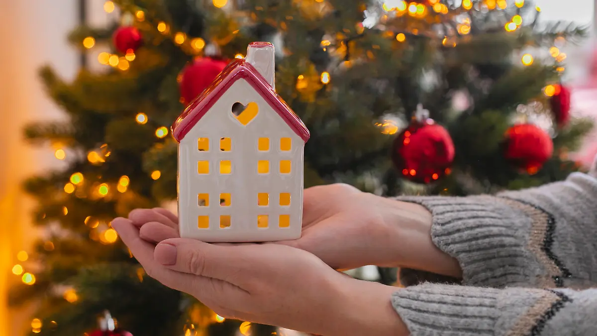 Zwei Hände halten ein Keramik-Lichthaus vor einem Weihnachtsbaum.