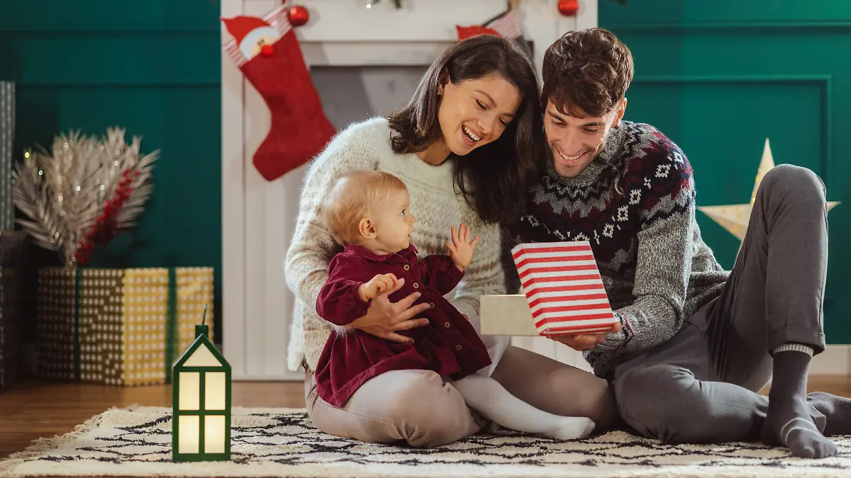 Junge Eltern sitzen mit Baby auf dem Boden und packen ein Weihnachtsgeschenk aus.