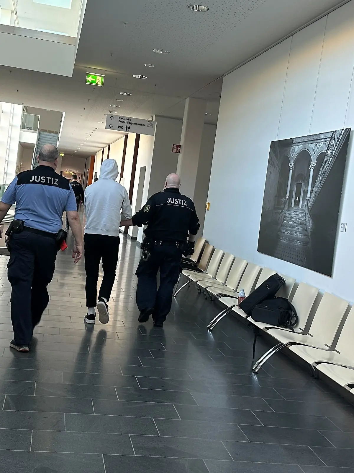 Ein Mann wird im Gericht von zwei Beamten flankiert und durch den Flur geführt. 