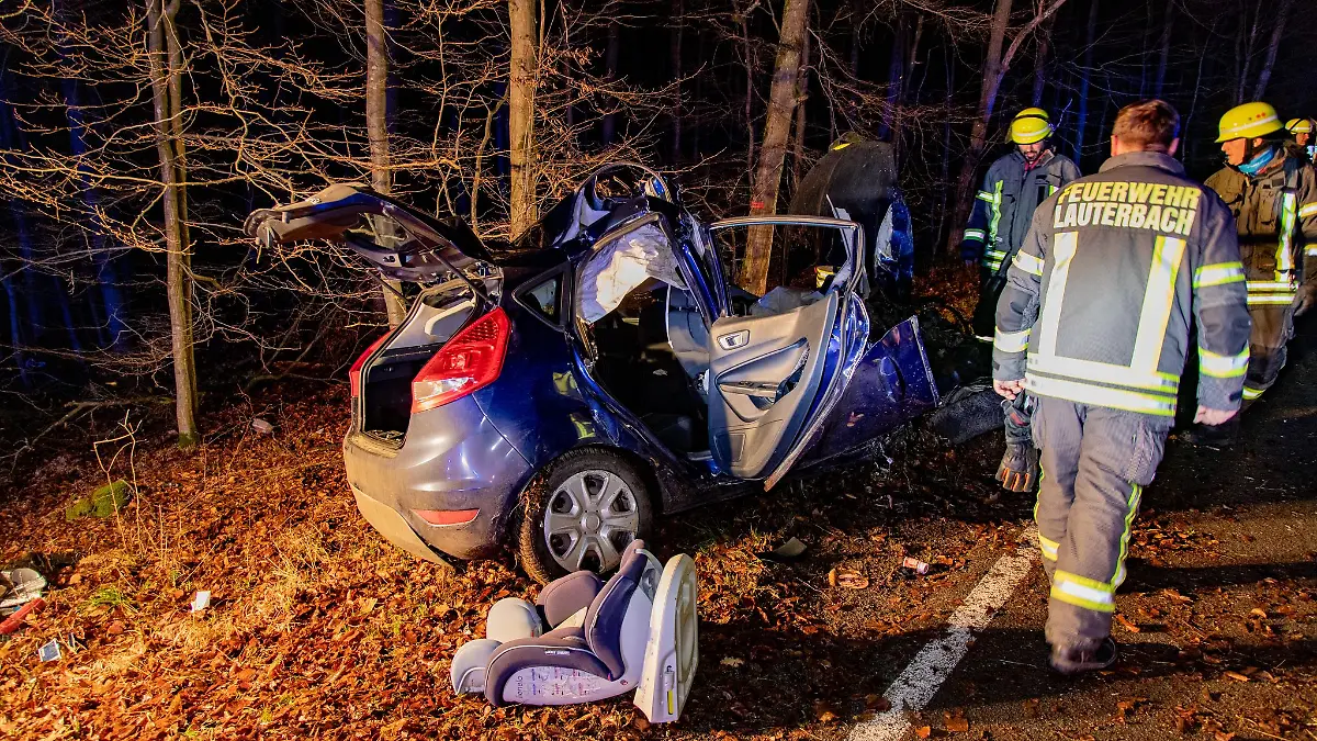 Feuerwehrmänner stehen neben einem verunglückten Auto. Ein mit vier jungen Menschen besetztes Auto ist im Vogelsbergkreis mit einem Baum zusammengestoßen und ein 17-jähriger Beifahrer gestorben.