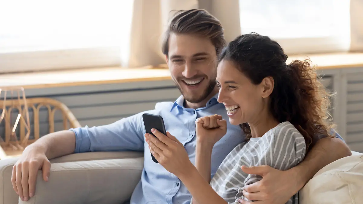 Happy young Caucasian couple relax on couch feel excited reading good news on smartphone, overjoyed man and woman rest on sofa triumph win online lottery on cellphone gadget, luck, reward concept
