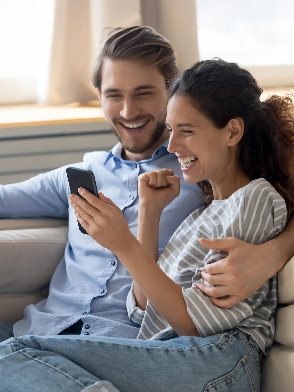 Happy young Caucasian couple relax on couch feel excited reading good news on smartphone, overjoyed man and woman rest on sofa triumph win online lottery on cellphone gadget, luck, reward concept