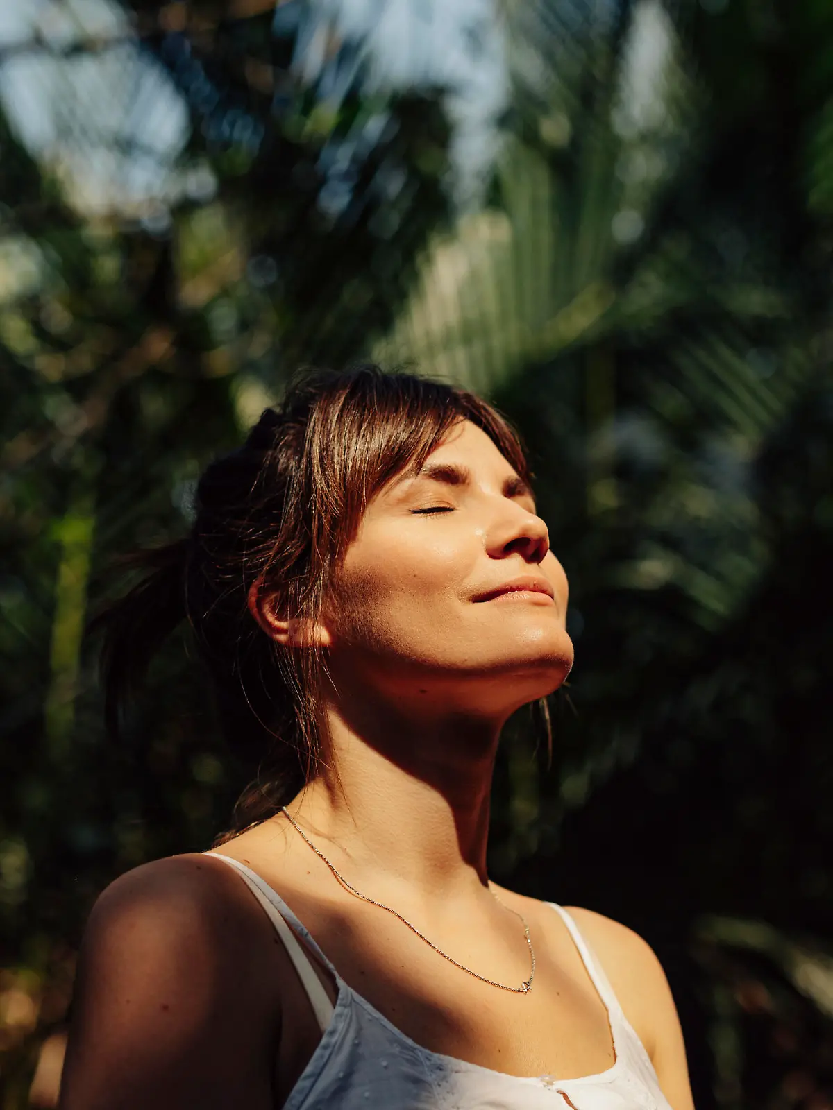 Close up upper body shot of a beautiful happy young Caucasian woman enjoying the warm sunlight and tropical atmosphere with her eyes closed surrounded by palm trees in a tropical public park.