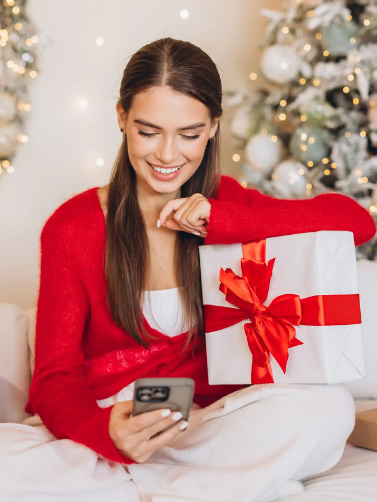 Eine Frau sitzt in weihnachtlicher Kulisse auf dem Boden und schaut auf ihr Smartphone.