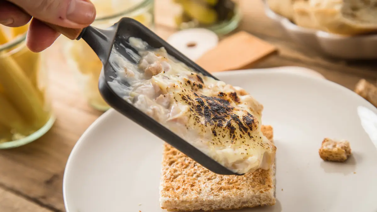 Von einem Raclette-Pfännchen läuft zerschmolzener Käse auf Brot.