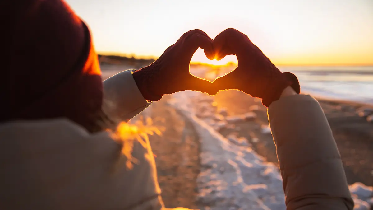 Eine Frau hält beim Winterspaziergang ihre Hände in die Luft und formt sie zu einem Herz. Im Hintergrund ist der Sonnenuntergang, die Landschaft ist voller Schnee.