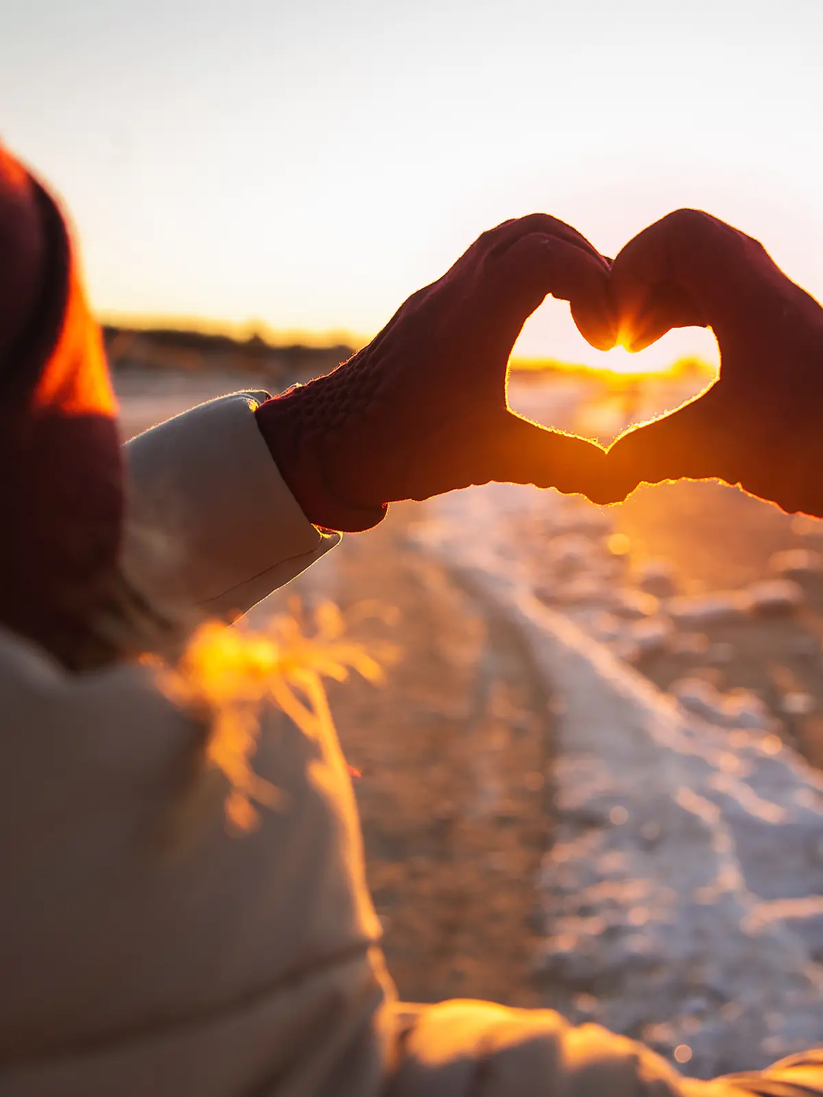 Eine Frau hält beim Winterspaziergang ihre Hände in die Luft und formt sie zu einem Herz. Im Hintergrund ist der Sonnenuntergang, die Landschaft ist voller Schnee.