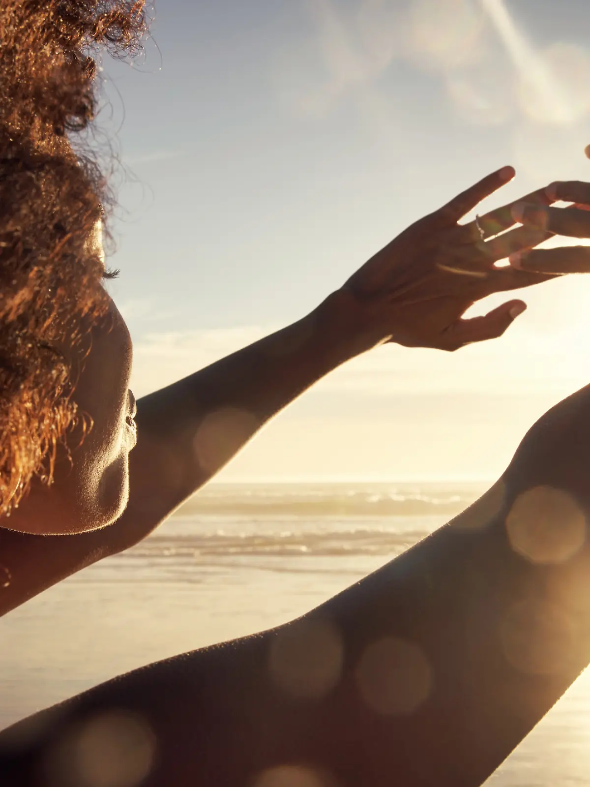 Eine Frau am Meer streckt ihre Arme zur Sonne hin.