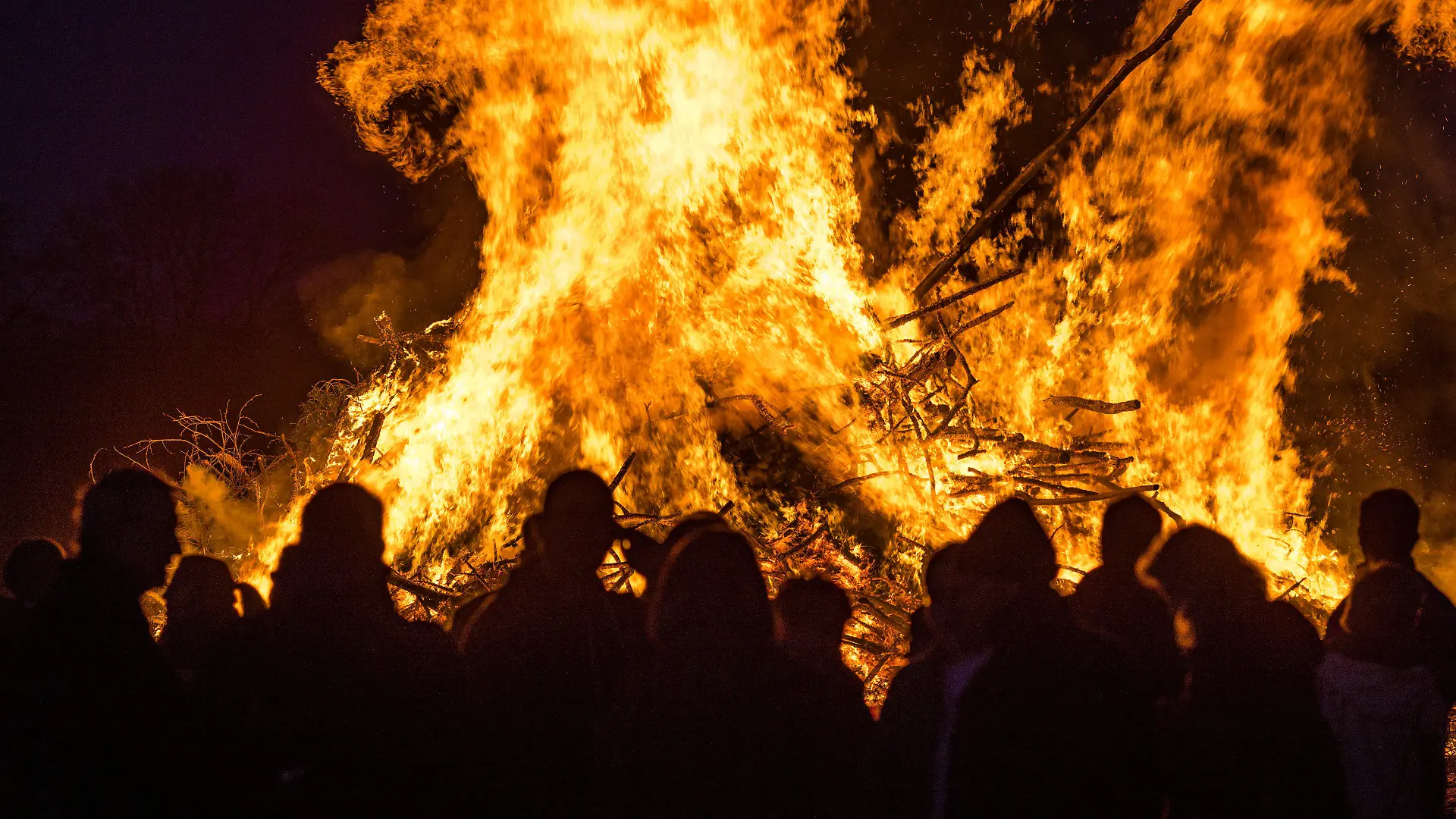 16.04.2022, Brandenburg, Cottbus: Besucher schauen sich das Branitzer Osterfeuer an. Die Kameraden der Freiwilligen Feuerwehr des Cottbuser Ortsteiles Branitz hatten bei den Einwohnern und Gewerbetreibenden etwa 75 Kubikmeter Holzabfall gesammelt und am Vormittag des heutigen Karsamstag zu einem eiförmigen Haufen geschichtet. Branitz hat zurzeit etwa 1500 Einwohner. Zu dem Osterfeuer kamen ungefähr 500 Besucher. Foto: Frank Hammerschmidt/dpa +++ dpa-Bildfunk +++