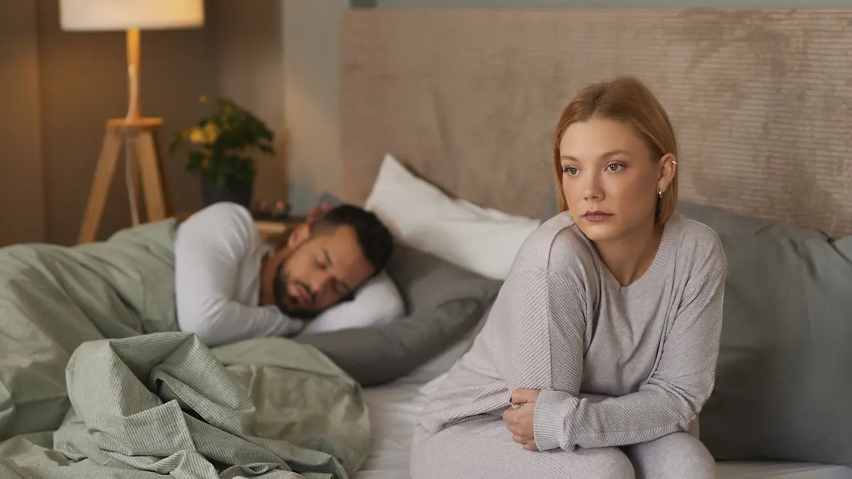 Eine nachdenkliche Frau sitzt auf der Bettkante, während ihr Partner im Hintergrund schläft.