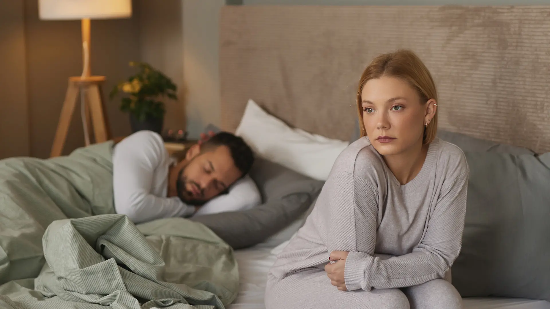 Eine nachdenkliche Frau sitzt auf der Bettkante, während ihr Partner im Hintergrund schläft.