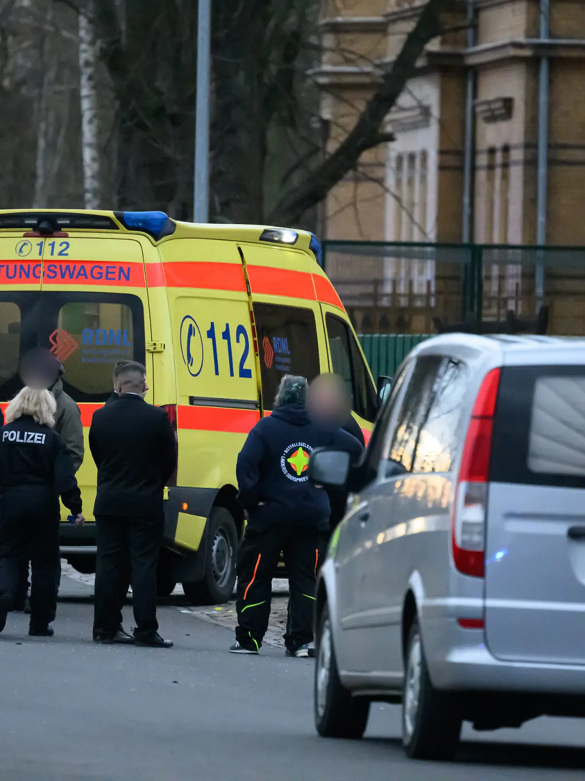 Polizisten stehen an einem Tatort vor einem Krankenwagen und einem Leichenwagen. Ein Polizist ist bei einer Fahndung nach Autodieben erfasst und getötet worden. 