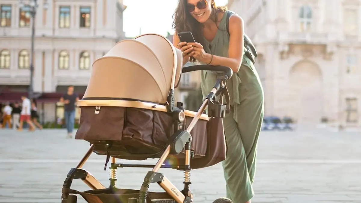 Frau mit sommerlichem Jumpsuit in hellgrün guckt lachend in ihren Kinderwagen und macht ein Foto mit dem Handy.