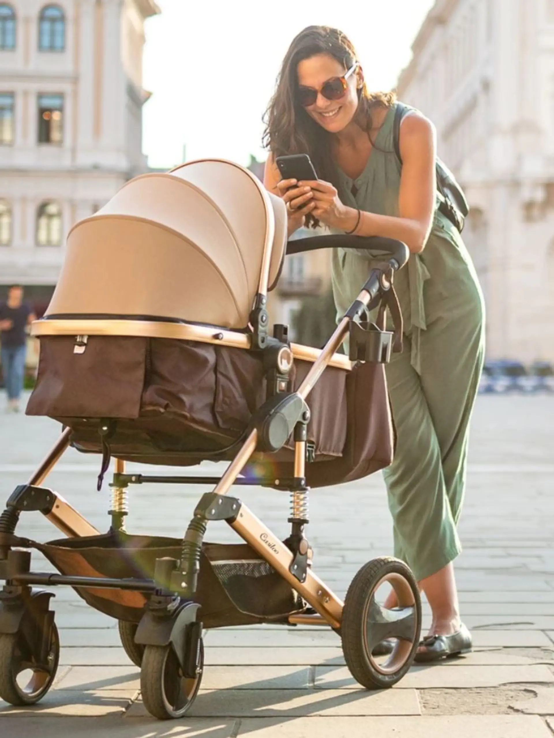 Frau mit sommerlichem Jumpsuit in hellgrün guckt lachend in ihren Kinderwagen und macht ein Foto mit dem Handy.