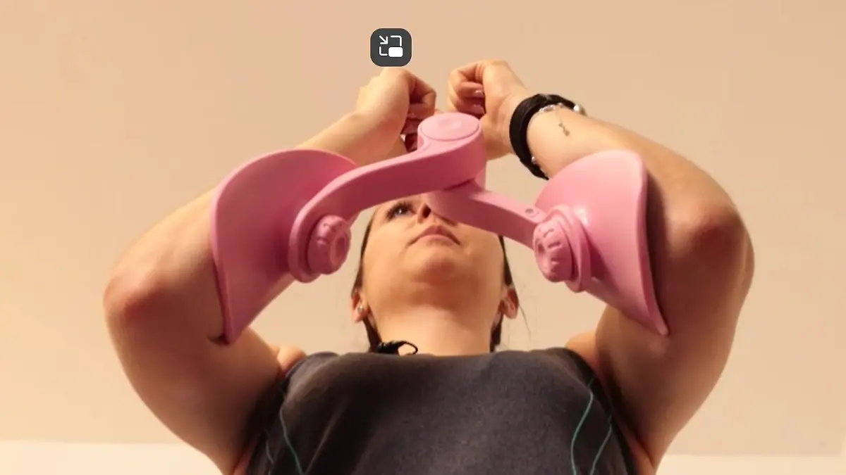 Frau macht mit einem rosafarbenen Fitnessgerät Armübungen, indem sie es zusammenpresst.