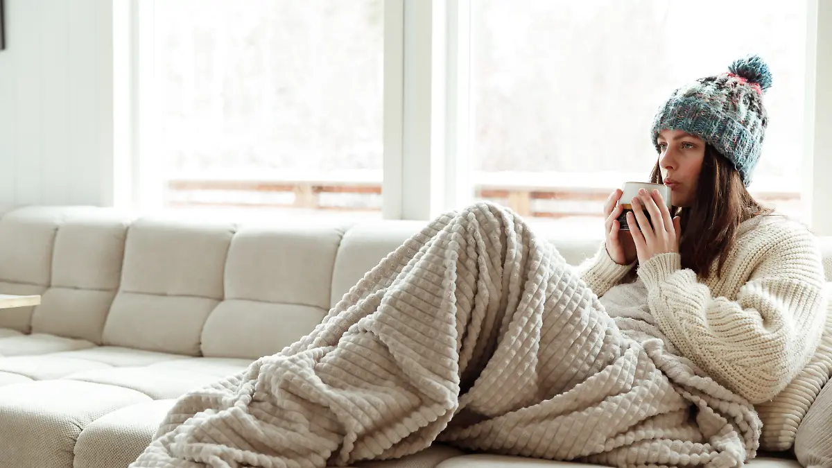 Frau sitzt mit Mütze und Decke in ihrem Wohnzimmer und friert