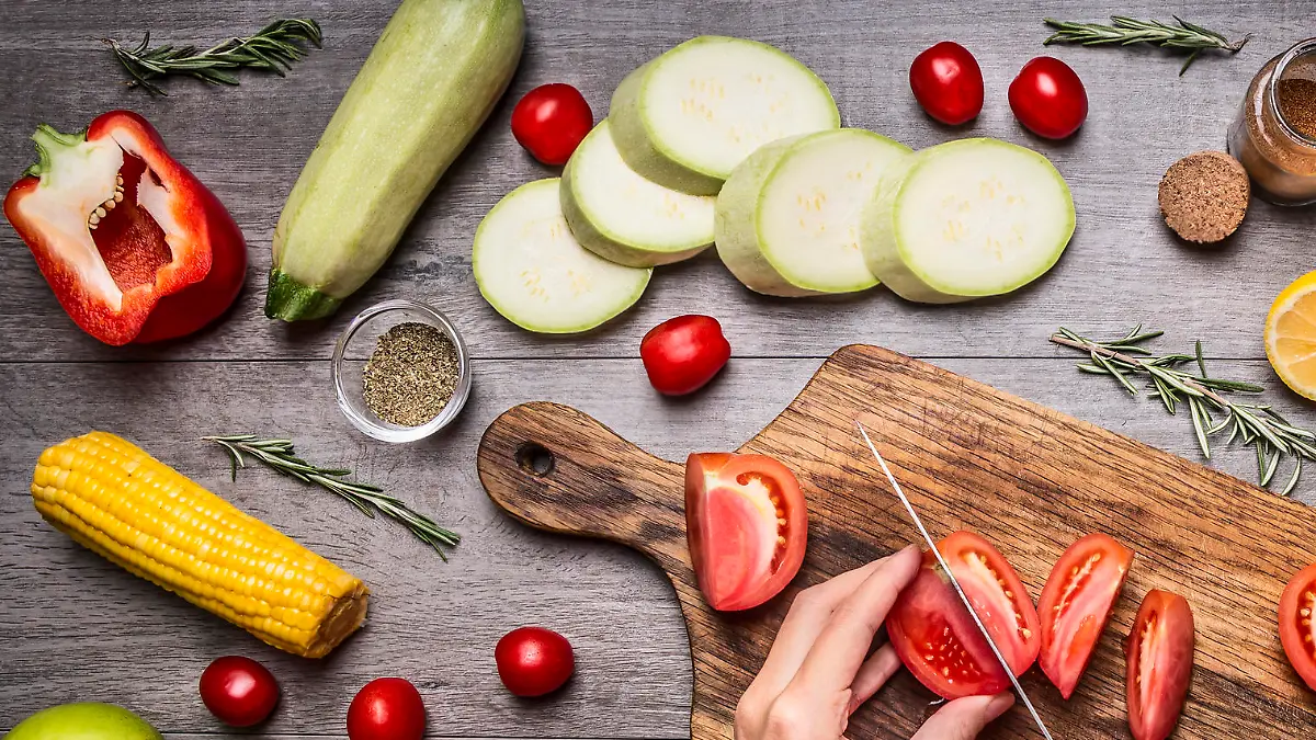 Female hand cut tomatoes on rustic kitchen table, around lie ingredients, vegetables, fruits, and spices, Healthy foods, cooking and vegetarian concept.