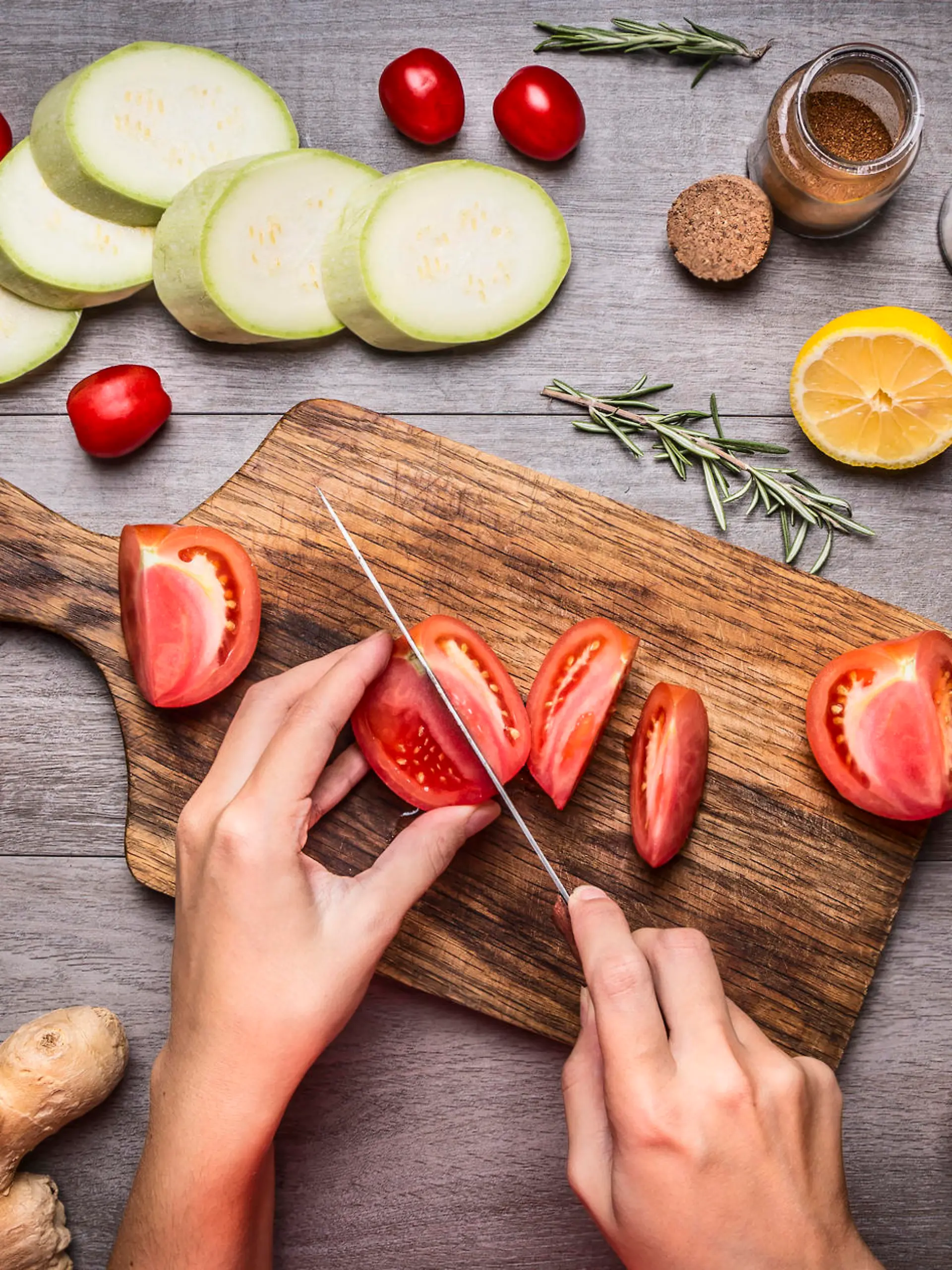 Female hand cut tomatoes on rustic kitchen table, around lie ingredients, vegetables, fruits, and spices, Healthy foods, cooking and vegetarian concept.